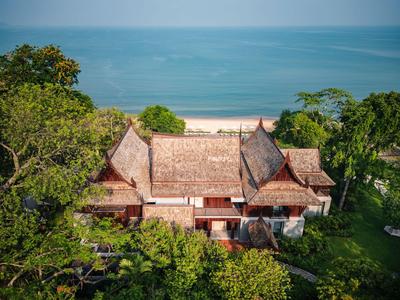 Traditional wooden house with thatched roof in lush greenery near a calm beach and sea.