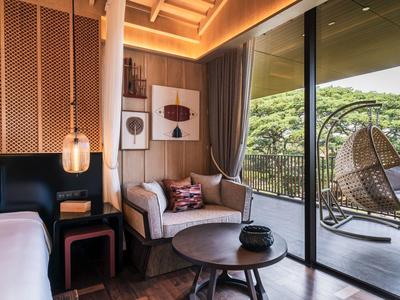 Modern hotel room with bed, sofa, coffee table, and hanging wicker chair on balcony with view.