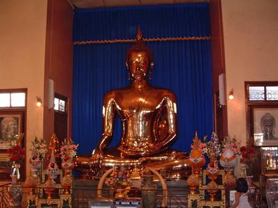 Statua di Buddha dorata seduta davanti a una tenda blu in un tempio circondata da composizioni di fiori.