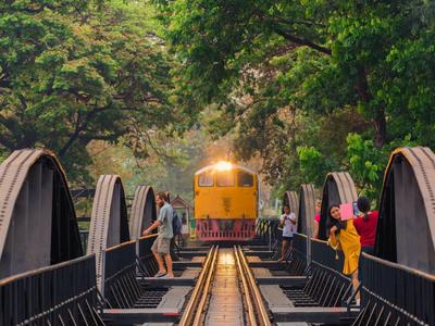 Yellow train crosses steel bridge with trees and tourists on sides.