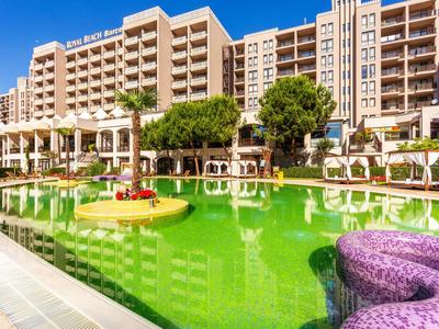 Modernes Hotel mit großem grünen Pool, Sonnenschirmen und mehrstöckigen Gebäuden im Hintergrund.