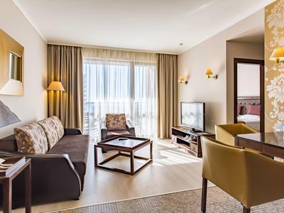 Helles Wohnzimmer mit Sofa, Sessel, Couchtisch, Fernseher und Zugang zu Balkon mit Gardinen.