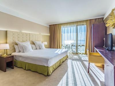 Helles Hotelzimmer mit Doppelbett, Nachttischen, TV und Balkon mit Ausblick.