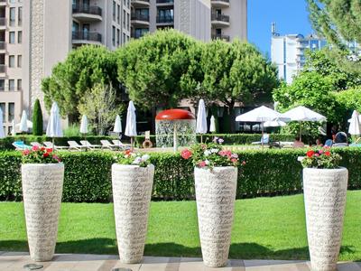 Vier große Pflanzgefäße mit blühenden Pflanzen vor einem Hotelgarten bei sonnigem Wetter.