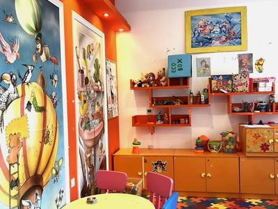 Bunter Kinderspielraum mit Tischen, Stühlen, Wandbildern und Regalen mit Spielzeug und Büchern.