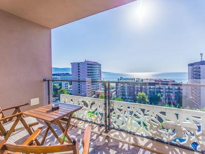 Balkon mit Holzstühlen und Tisch, Blick auf Stadt und Berge bei klarem Himmel.