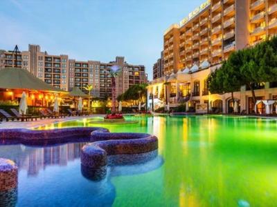 Beleuchteter Hotelpool bei Sonnenuntergang mit umliegenden Gebäuden und Außenrestaurants.