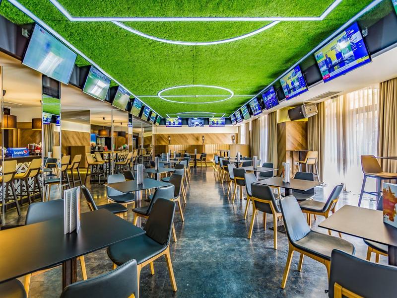 Moderner, gut beleuchteter Raum mit Tischen, Stühlen und Bildschirmen an den Wänden für Sportübertragungen.