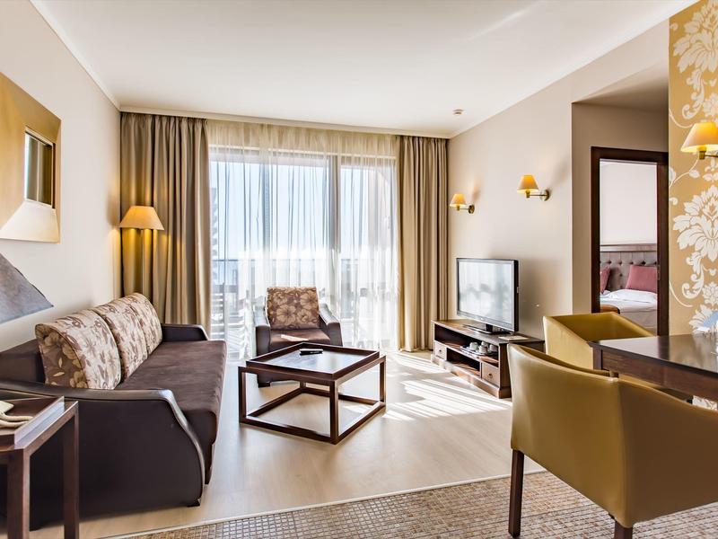 Helles Wohnzimmer mit Sofa, Sessel, Couchtisch, Fernseher und Zugang zu Balkon mit Gardinen.