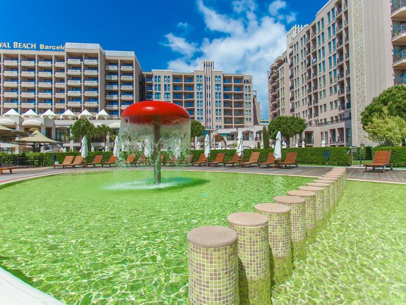 Outdoor-Pool mit Pilz-Wasserspiel vor Hotelgebäuden bei sonnigem Himmel