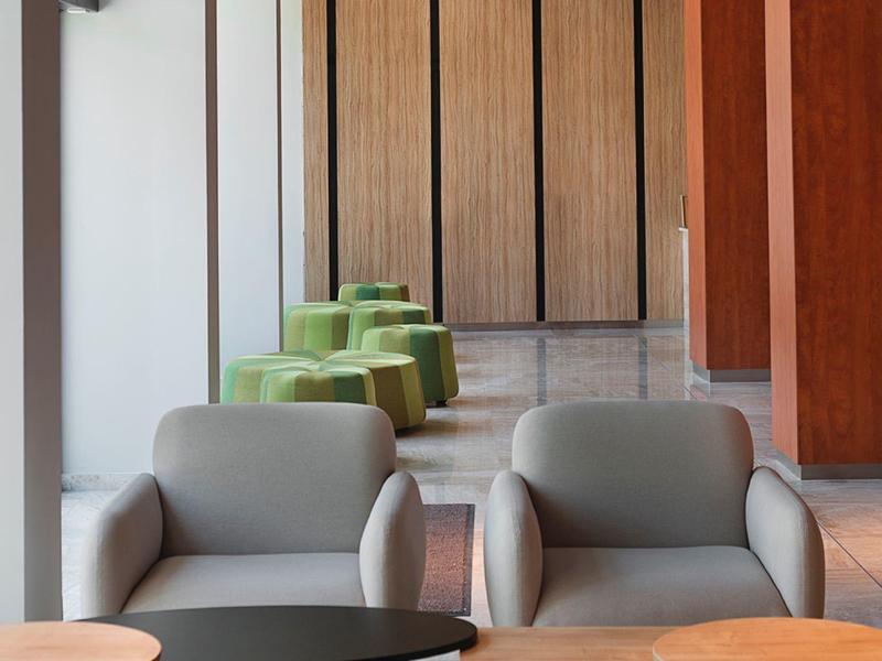 Moderní hotelová hala se šedými křesly, dřevěnými stolky a zelenými stoličkami před dřevěnou stěnou.