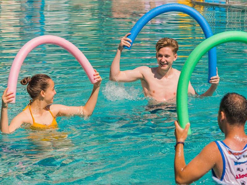 Tři lidé v hotelovém bazénu si hrají s barevnými bazénovými tyčemi a smějí se.