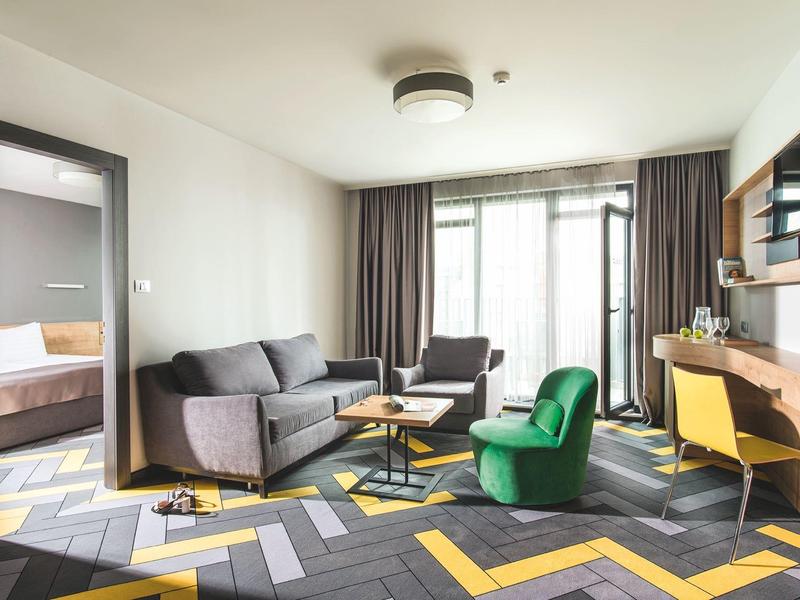 Moderně zařízený hotelový pokoj s posezením, pohovkou, zeleným křeslem a psacím stolem.