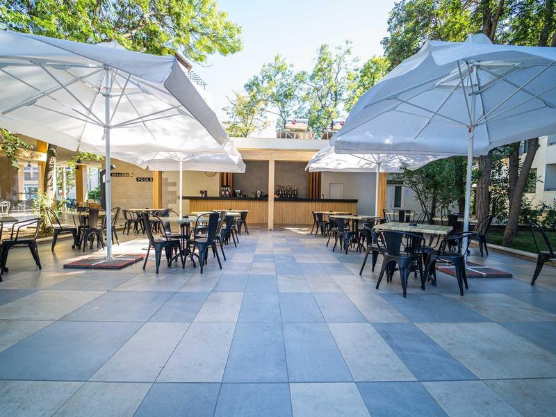 Velká terasa s ​​stoly, židlemi a slunečníky v hotelové zahradě pod stromy.