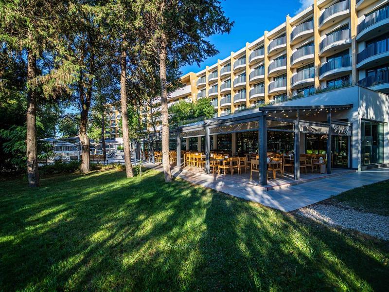 Moderní hotelová budova s otevřenou restaurací a zastíněnou zahradou v popředí.