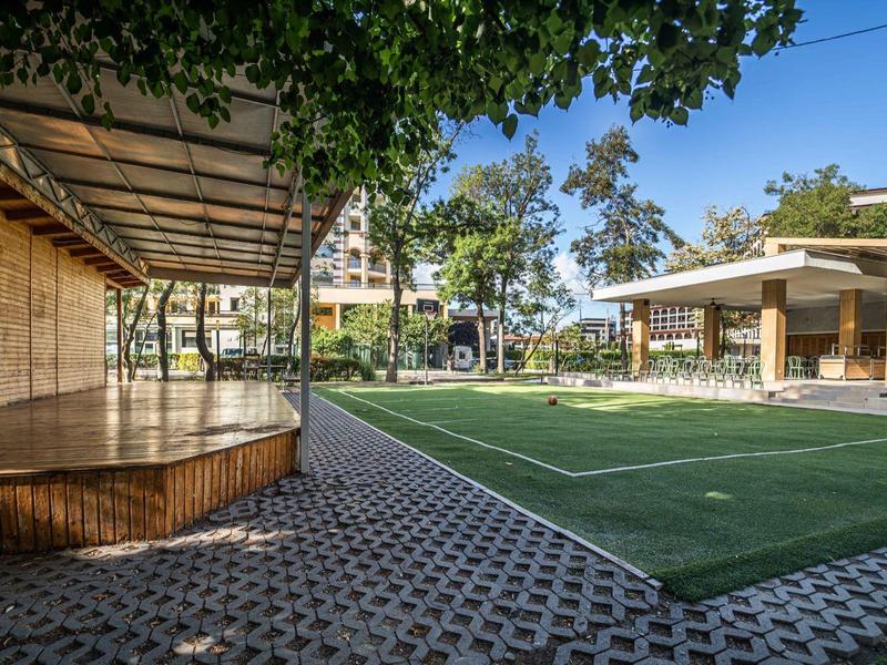 Moderní venkovní prostor s krytým posezením a umělým trávníkem pod stromy.