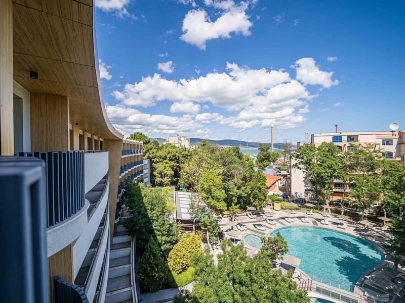 Výhled z okna hotelu na bazén obklopený stromy a budovami pod modrou oblohou.