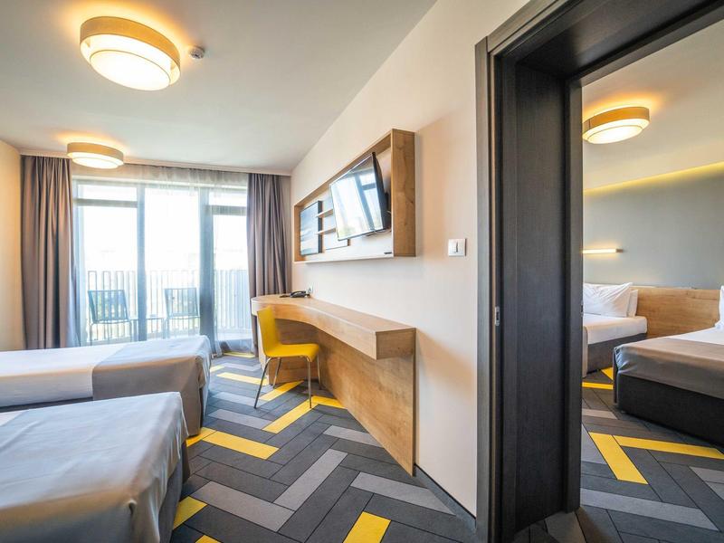 Moderní hotelový pokoj se dvěma postelemi, žlutými akcenty a balkonem s výhledem.