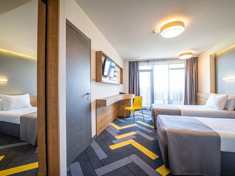 Moderní hotelový pokoj se dvěma jednolůžky, stolem, televizí a velkým oknem se závěsy.