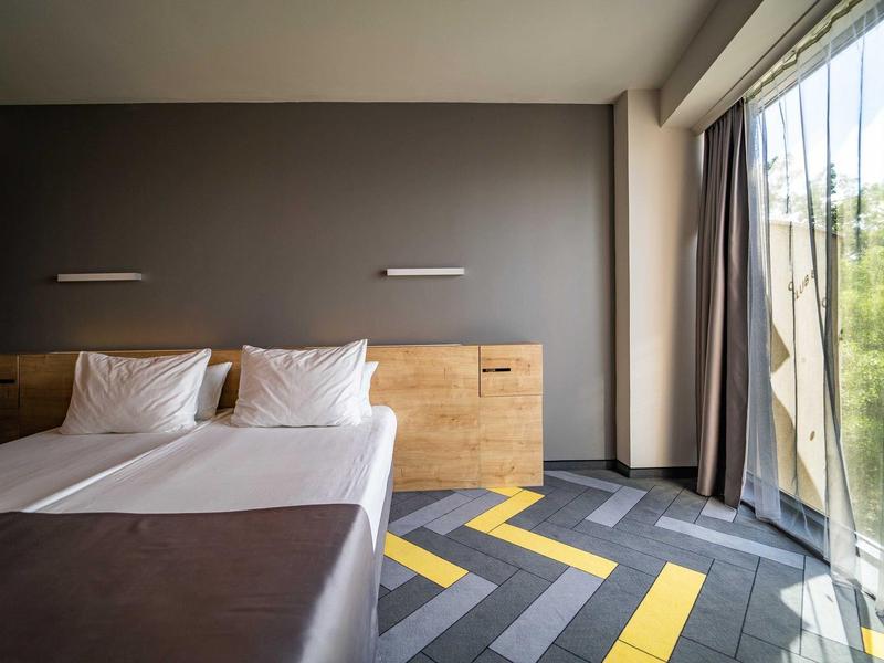 Moderní hotelový pokoj s dvojlůžkem, dřevěnými čely postele a velkými okny s výhledem na zeleň.