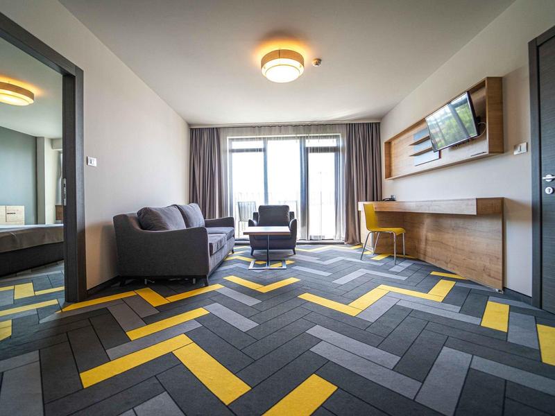 Moderně zařízený hotelový pokoj s pohovkou, křeslem, televizí a barevným kobercem.