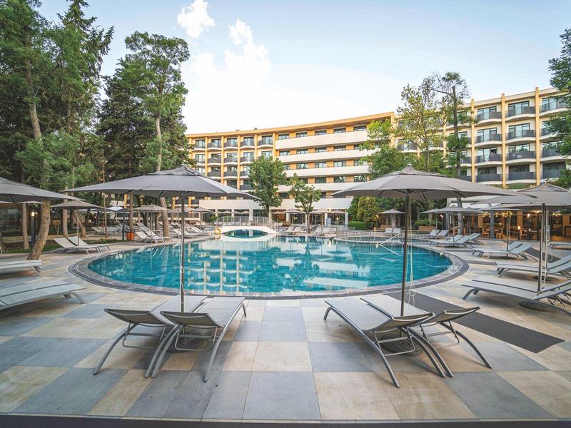 Kruhový bazén s lehátky a slunečníky před vícepodlažní hotelovou budovou.