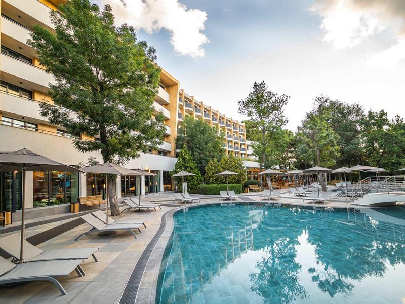 Hotel s velkým venkovním bazénem, lehátky a posezením pod slunečníky za zataženého dne.