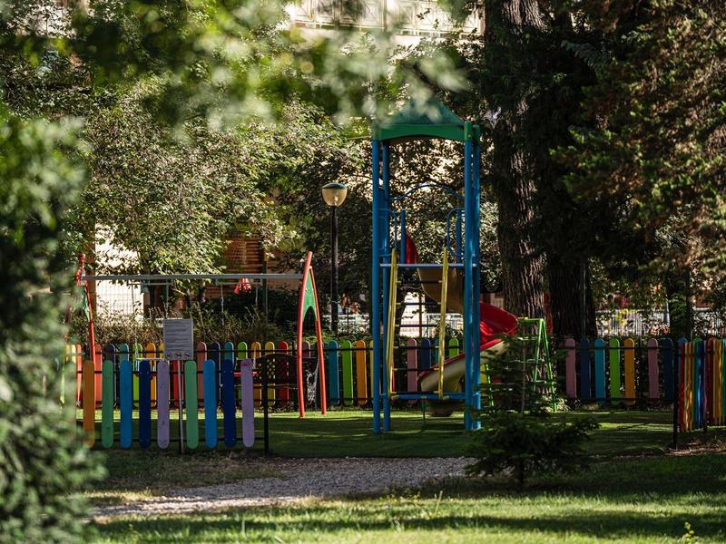 Barevné dětské hřiště s houpačkami a plotem v zalesněném parku.