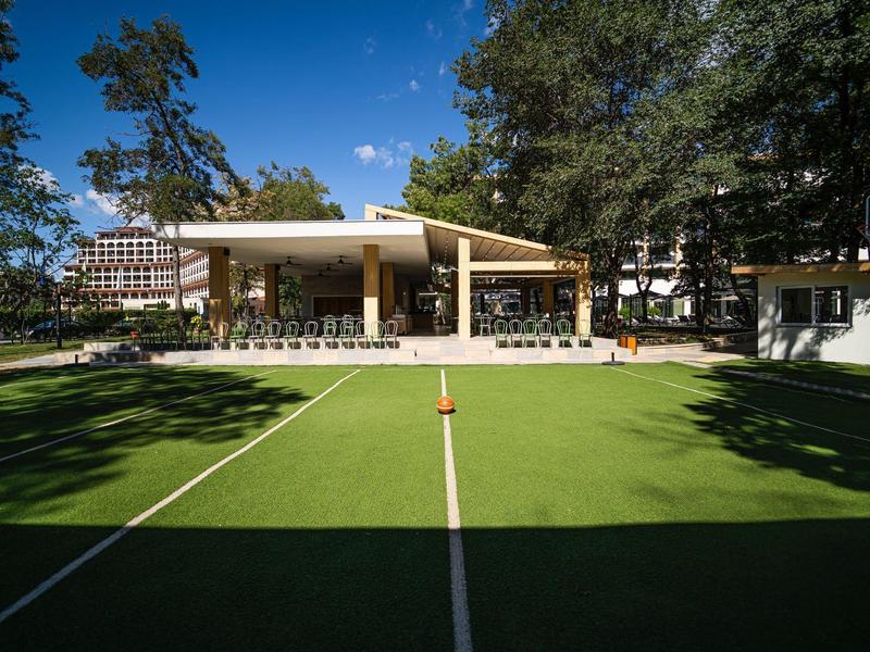 Pohled na udržované tenisové hřiště s moderními budovami a stromy pod modrou oblohou.