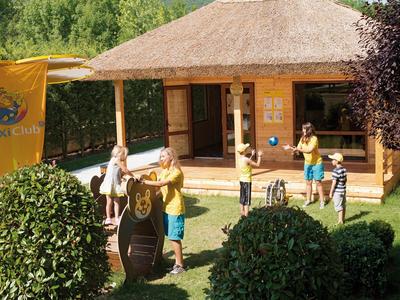 Menschen spielen Minigolf vor einem Holzhaus mit Strohdach, umgeben von grünen Büschen und Bäumen.