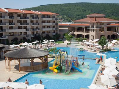 Großer Hotelpool mit buntem Kinderspielplatz, Sonnenliegen und umliegenden Gebäuden vor bewaldetem Hügel.