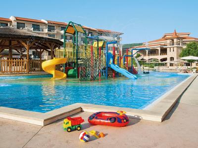 Kinderpool mit bunten Rutschen, Spielgeräten und Spielzeugautos vor Hotelgebäuden an sonnigem Tag.