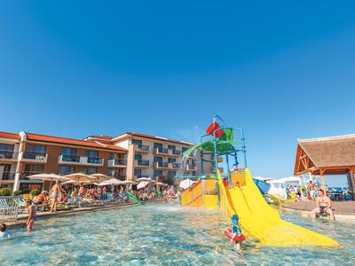 Großes Schwimmbad mit gelber Rutsche, Spielgeräten und umgeben von Hotelgebäuden unter blauem Himmel.