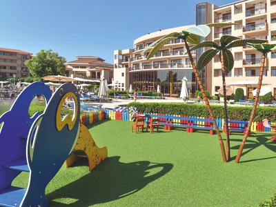 Bunter Kinderspielplatz mit Rutsche, Grasboden und zwei großen Palmen aus Kunststoff vor Gebäuden.