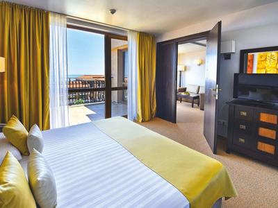 Modernes Hotelzimmer mit großem Bett, gelben Vorhängen und Blick auf eine Terrasse mit Meer.