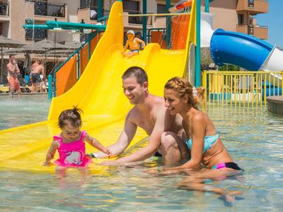 Familie spielt im flachen Wasser an einer gelben Rutsche im Schwimmbad bei Sonnenschein.