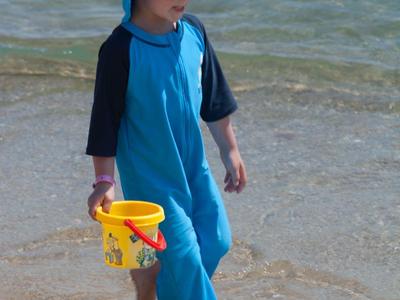 Kleines Kind im blauen Sonnenschutzanzug geht mit gelbem Eimer am Strand ans Wasser.