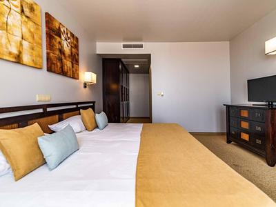 Hell eingerichtetes Hotelzimmer mit Doppelbett, braunem Bettüberwurf und Fernseher auf Kommode.