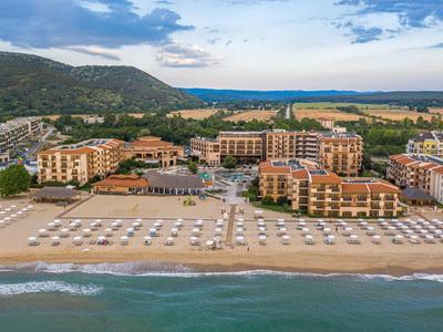 Breiter Sandstrand mit Reihen weißer Sonnenschirme vor mehreren mehrstöckigen Hotelgebäuden und grünen Hügeln.