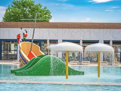 Buntes Wasserspiel mit Rutsche, Wasserpilzen und grünem Hügel im Schwimmbad bei blauem Himmel.