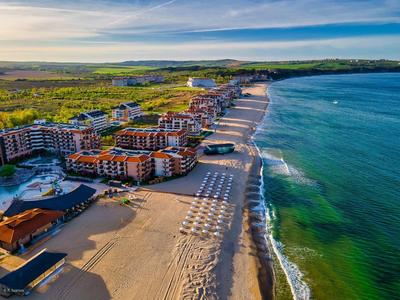 Luftaufnahme eines Strandes mit Hotels, Sonnenschirmen und grünem Meer unter blauem Himmel.