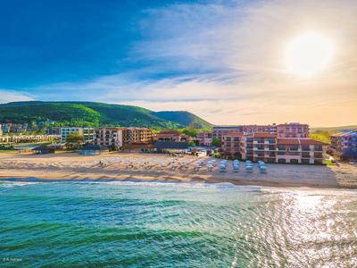 Strand mit grün bewachsenen Hügeln, mehreren Gebäuden und hellem Sonnenlicht am klaren Himmel.