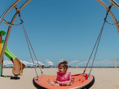 Kind sitzt auf einer runden Schaukel am sandigen Strand bei klarem Himmel.