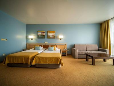 Hotelzimmer mit zwei Einzelbetten, Sofa, Holzmöbeln und großen Fenstern mit Vorhängen.