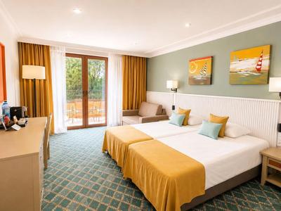 Hell eingerichtetes Hotelzimmer mit Doppelbett, großen Fenstern und Zugang zu einem Balkon.