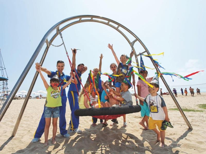 Niños juegan en un área de juegos estilo caribeño frente a un edificio con techo de paja en el resort.