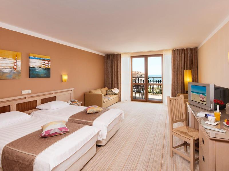 Habitación de hotel luminosa con dos camas individuales, sofá, televisor y balcón con vista al mar.