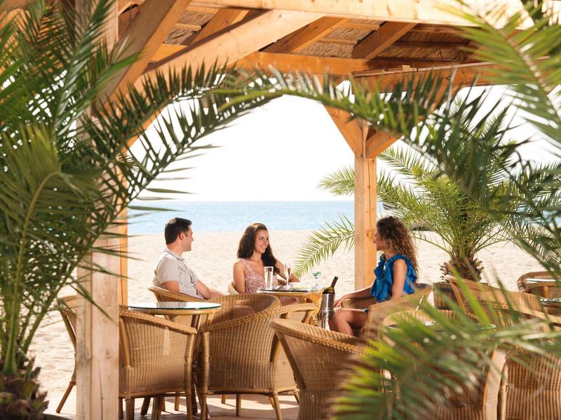 Tres personas conversan en una terraza de madera con vista al mar y plantas verdes.