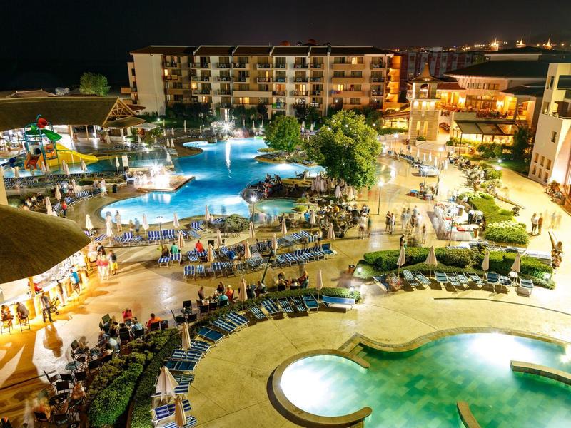 Vista nocturna de piscina hotelera iluminada con tumbonas y huéspedes.