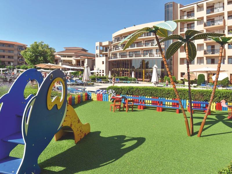 Área de juegos colorida con tobogán y palmeras frente a edificios de hotel bajo el sol.